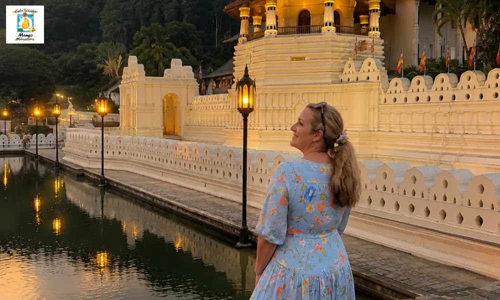 A traveler stands beside the tranquil lake in front of the Temple of the Sacred Tooth Relic in Kandy, Sri Lanka, admiring the beautifully illuminated temple at dusk with its reflection shimmering on the water.