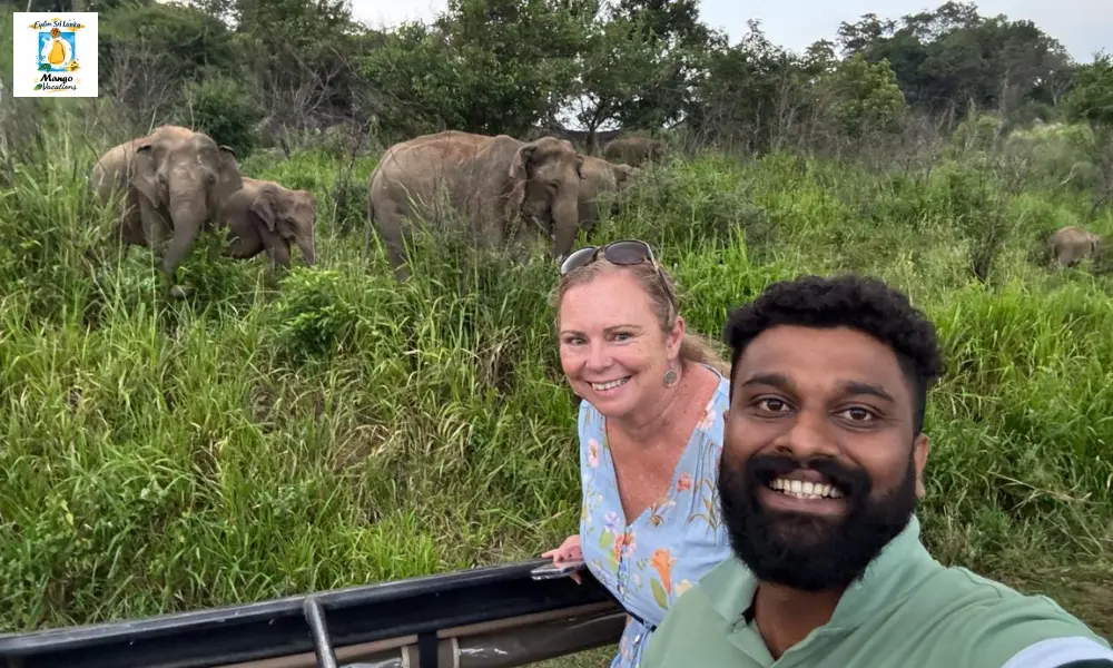 A traveler and a Mango Vacations guide smile for a selfie during a safari in Yala National Park, Sri Lanka, with a herd of wild elephants grazing peacefully in the background.