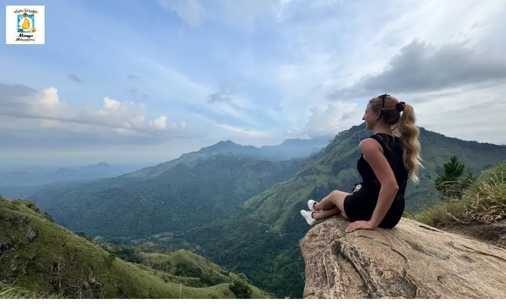 A traveler sits on a rocky ledge at Little Adam's Peak in Ella, Sri Lanka, overlooking rolling green mountains and lush valleys beneath a dramatic blue sky with scattered clouds.