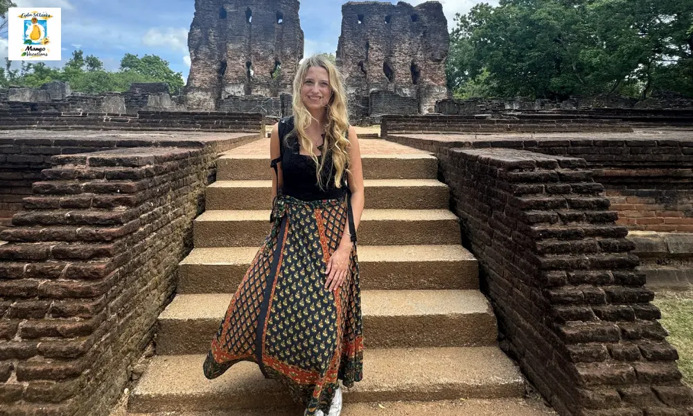 A female traveler stands on the stone steps of the ancient Royal Palace ruins in Polonnaruwa, Sri Lanka, smiling at the camera while surrounded by centuries-old brick architecture and lush green trees.